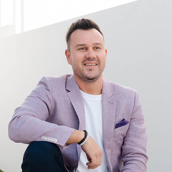 Smiling man in light purple blazer and white shirt, Gold Coast mortgage adviser, sits outdoors by a white wall.