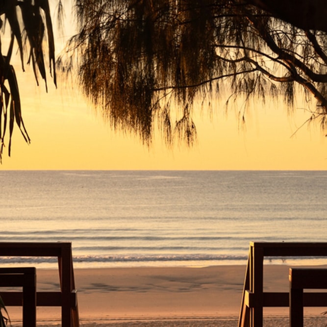Sunset at a tranquil Gold Coast beach with calm waves, golden water, trees and wooden chairs—ideal for a Mortgage Adviser Gold Coast.
