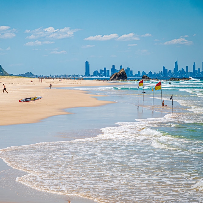 Sandy beach with gentle waves, surfboard, and city skyline—relaxing scene near Mortgage Broker Gold Coast.