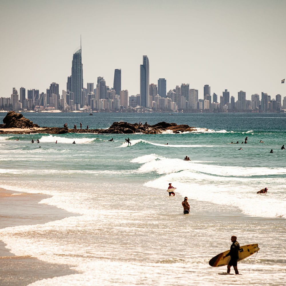 Swimmers and surfers enjoy gentle waves near a rocky outcrop with Gold Coast skyscrapers in view; Mortgage Broker Gold Coast.