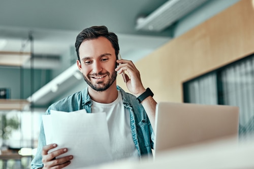 A smiling Mortgage Broker Gold Coast talks on his mobile, holding papers and sitting at a laptop in a modern office.